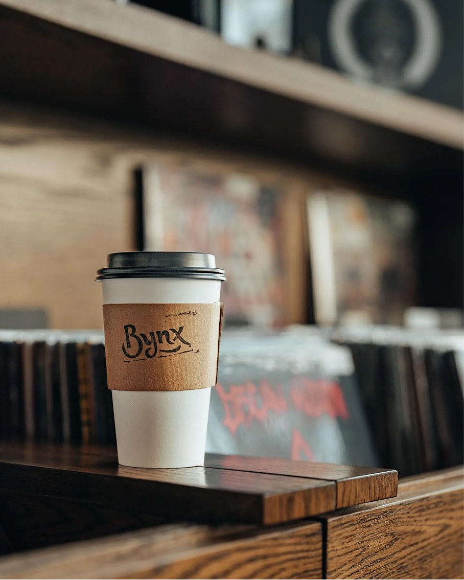 A Bynx to-go coffee cup with a stamped sleeve in front of shelves of vinyl records