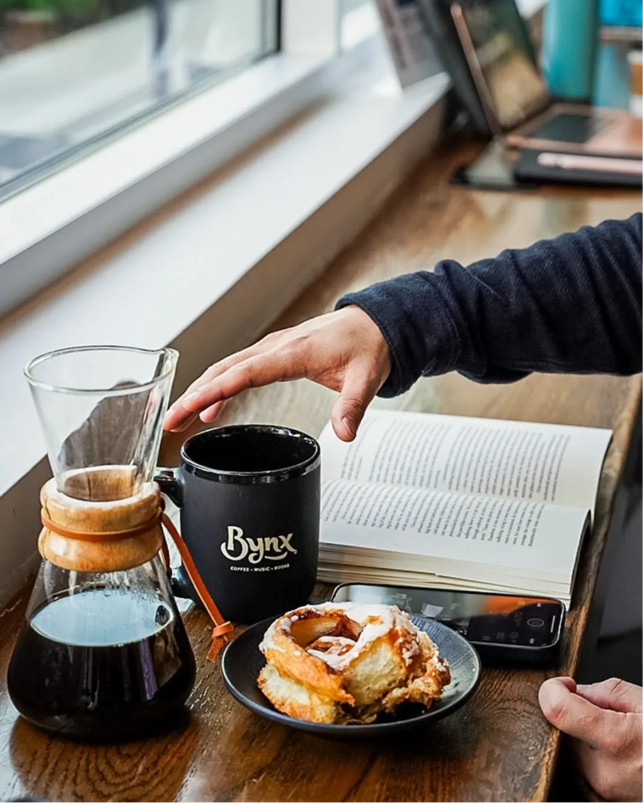 A Bynx mug, pour-over coffee, pastry, and open book on a windowsill counter